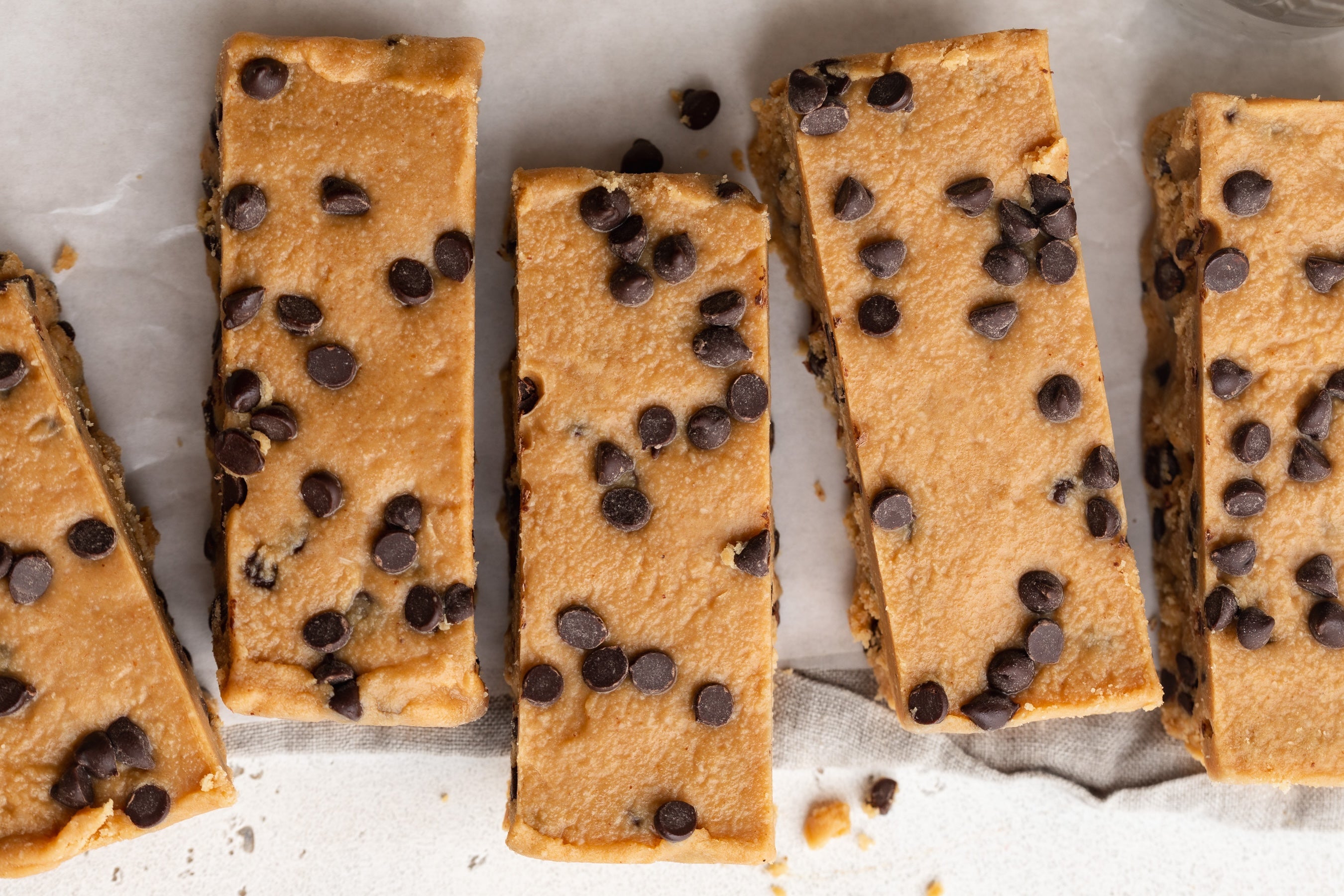 Chocolate chip cookie bars arranged on parchment paper, showing a golden-brown cookie base with scattered dark chocolate chips.