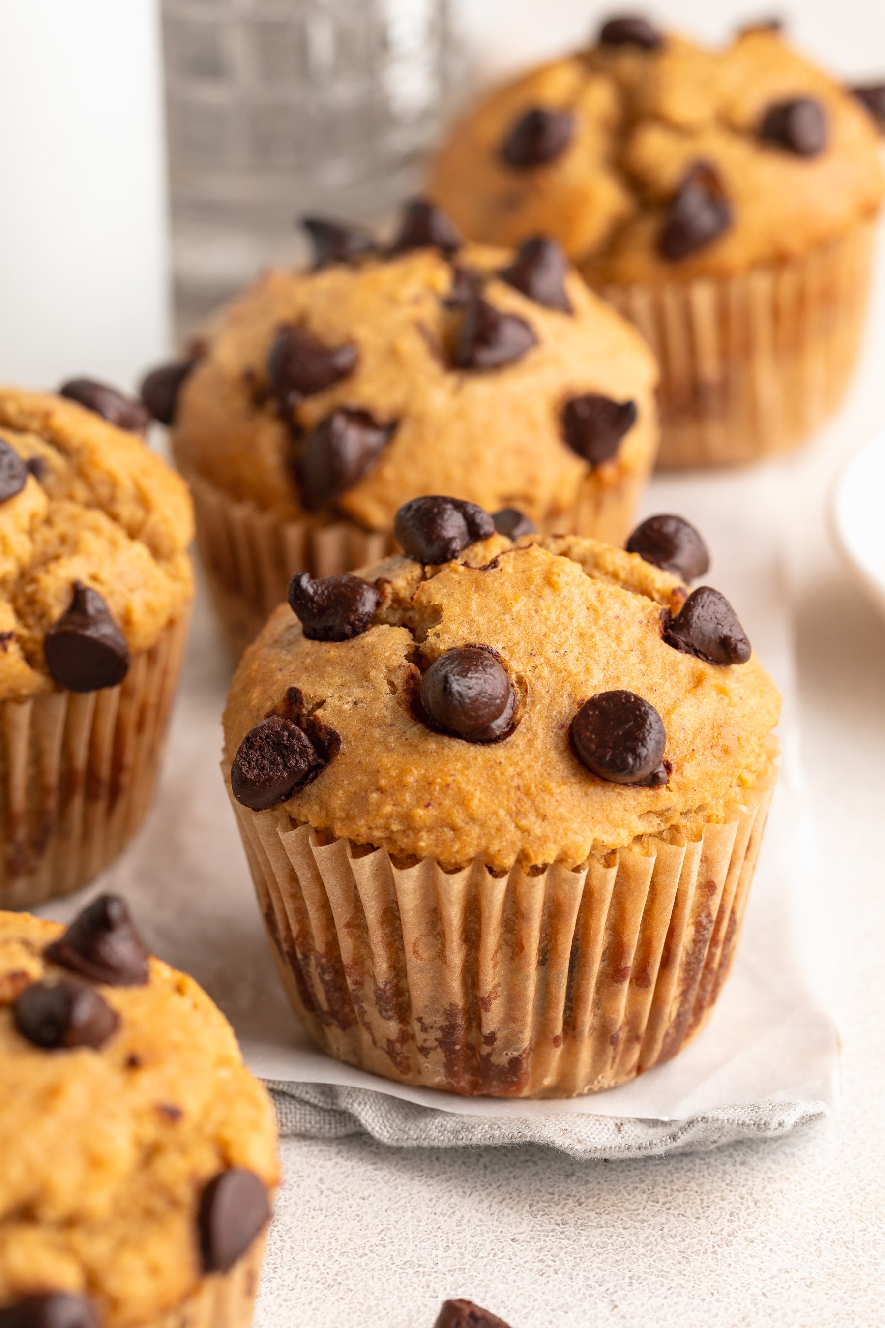 Chocolate chip muffins in paper liners, with one in the foreground and several more blurred in the background on a light surface.