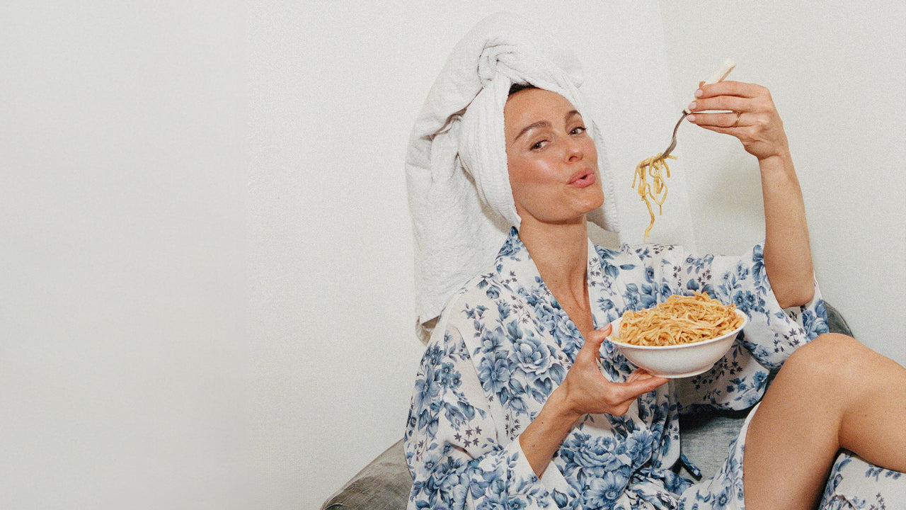 Megan in a floral robe and towel on her head eating noodles from a bowl.