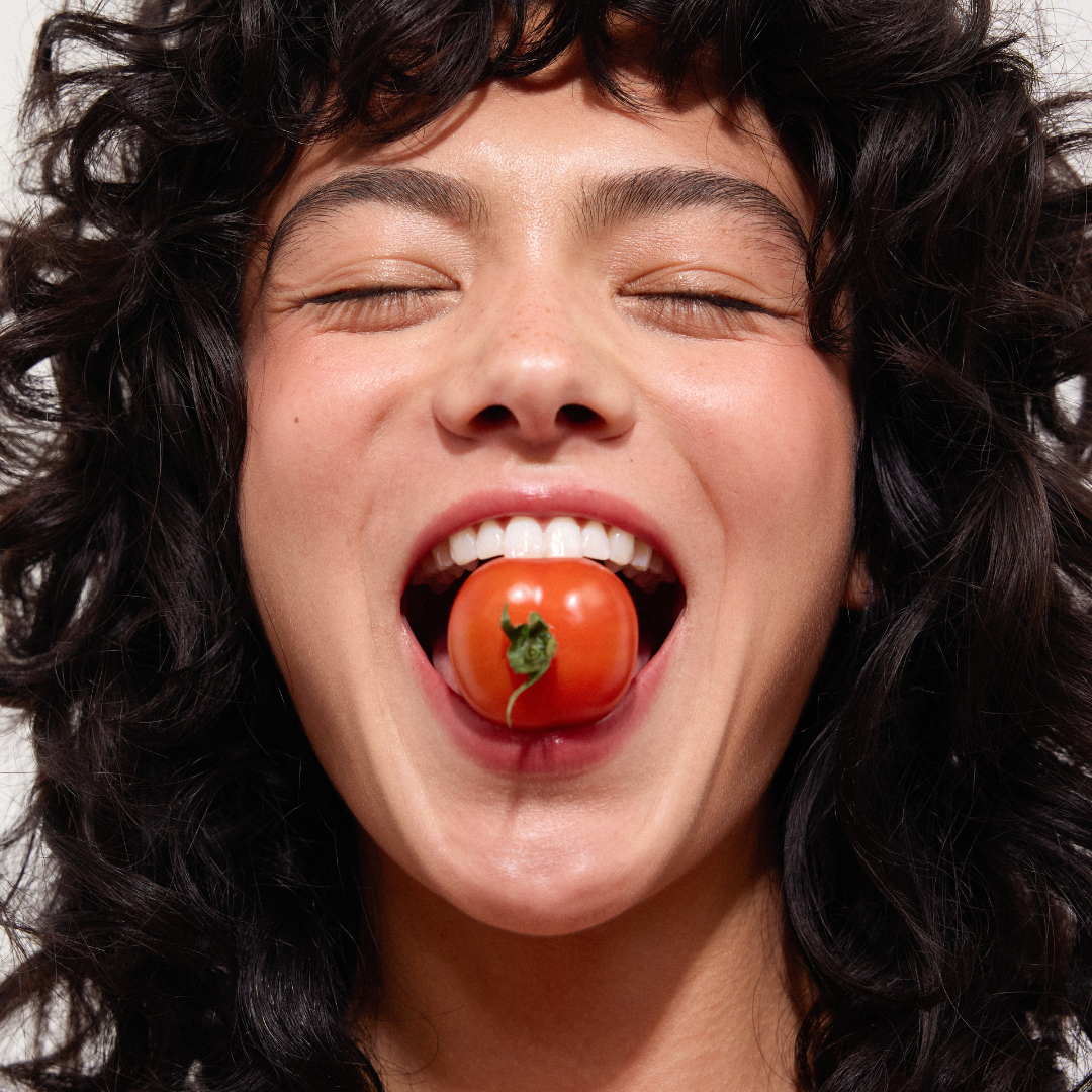 Woman with dark hair smiling with a tomato in her mouth