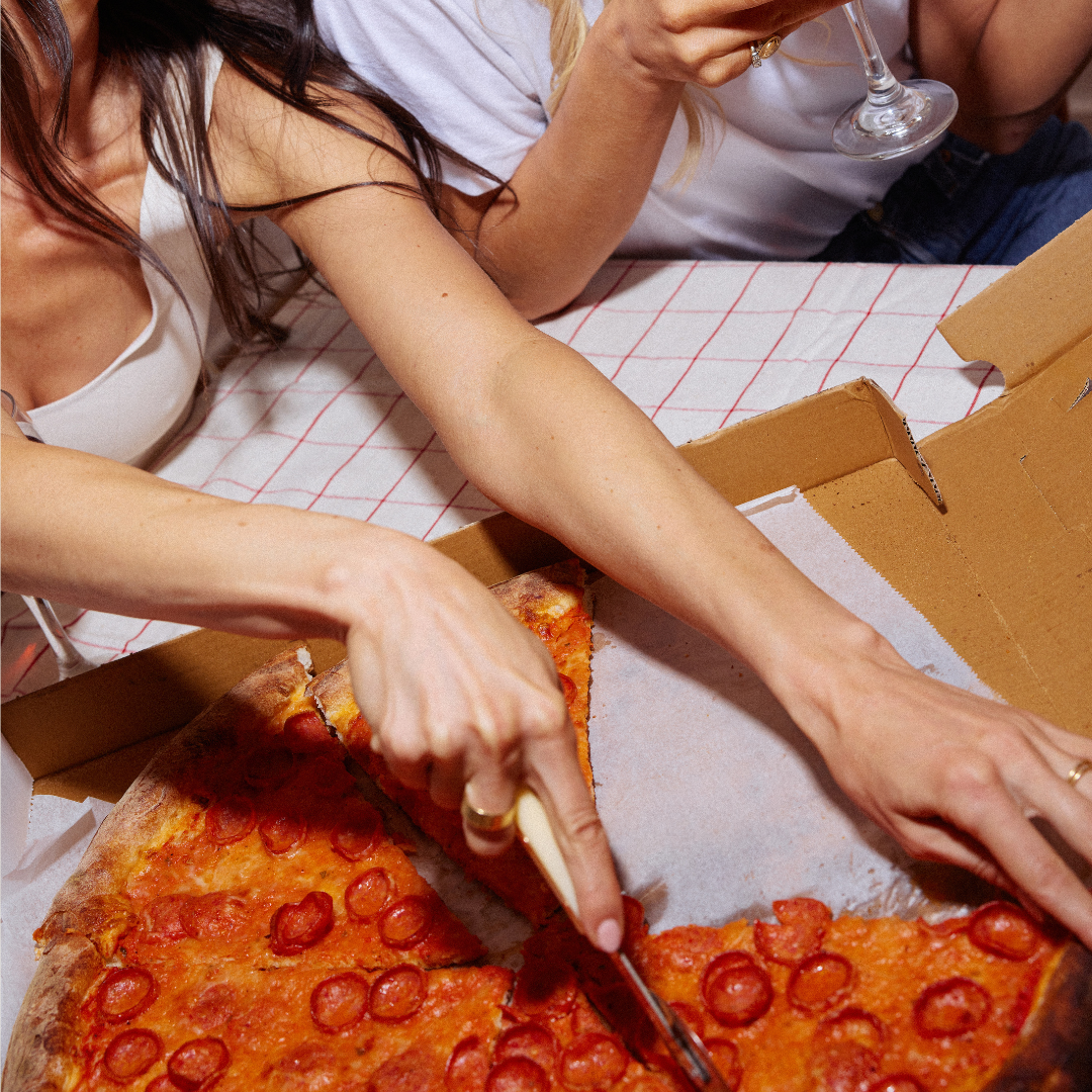 Person cutting a pepperoni pizza with a knife on a checkered tablecloth.