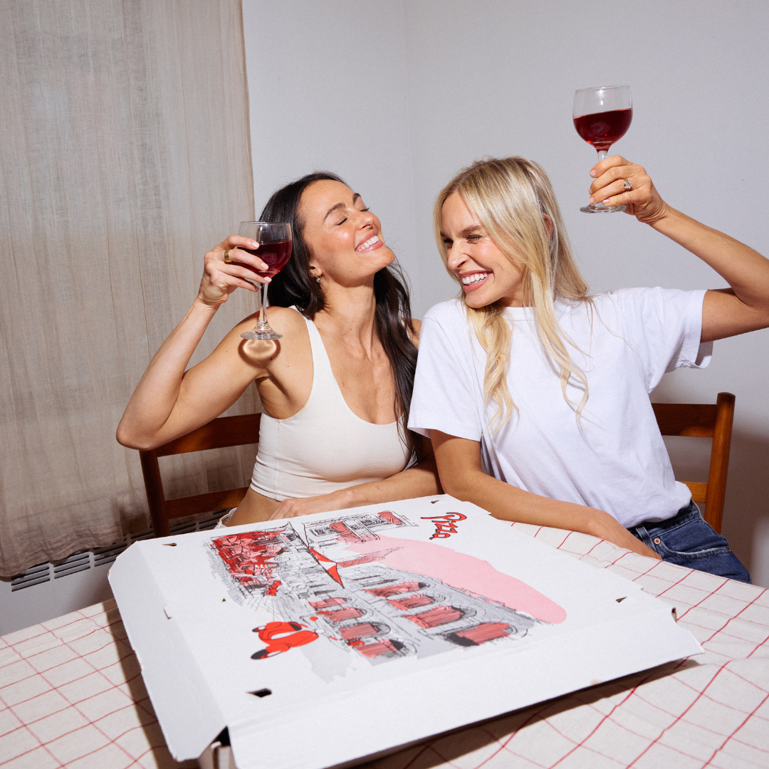 Megan and Katy sitting at a table with a pizza box, raising wine glasses.