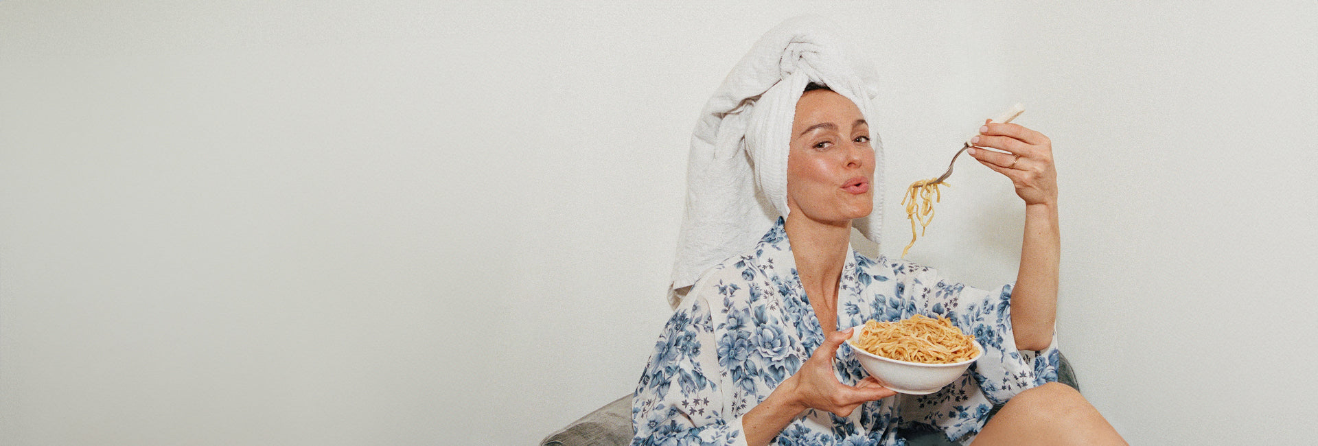 Woman in a floral robe eating pasta with a towel on her head against a white background