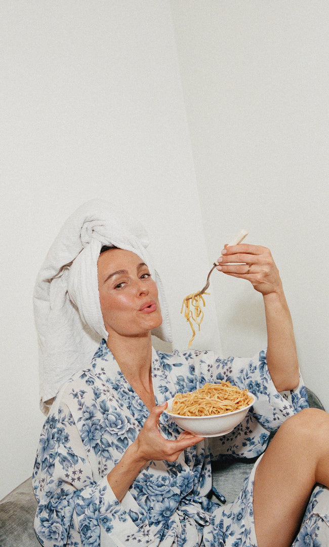 Woman in a floral robe eating spaghetti with a towel on her head against a white background